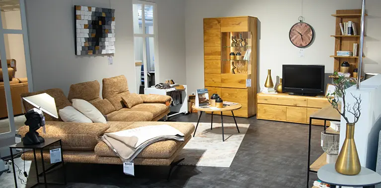 Sitzgruppe mit braunem Sofa, Couchtisch und Wohnwand aus Holzoptik, ergänzt durch Regal und Wandbild in neutralem Ausstellungsraum.